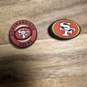 On a wooden surface, there are two Football silicone bead San Francisco coasters. One is circular and red, featuring the team logo and name, while the other is oval-shaped with an alternate version of the logo.