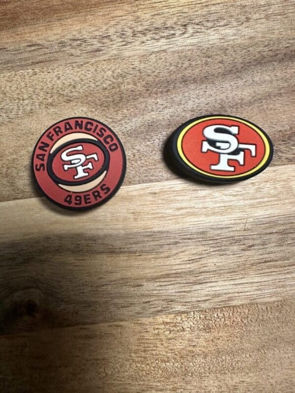 On a wooden surface, there are two Football silicone bead San Francisco coasters. One is circular and red, featuring the team logo and name, while the other is oval-shaped with an alternate version of the logo.