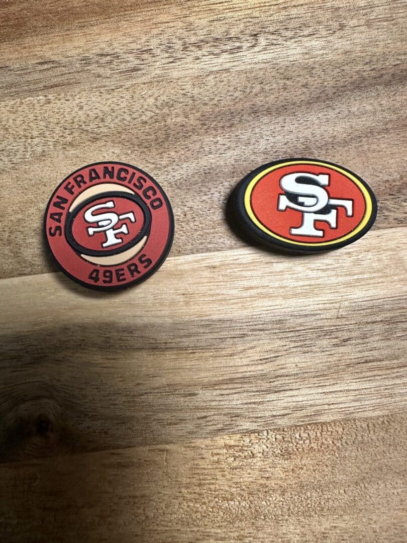 On a wooden surface, there are two Football silicone bead San Francisco coasters. One is circular and red, featuring the team logo and name, while the other is oval-shaped with an alternate version of the logo.