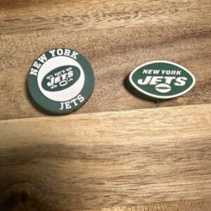 Two New York Jets accessories on a wooden surface. On the left is a circular NY football silicone focal bead with the team logo, and on the right is an oval-shaped NY football silicone focal bead with the same logo.