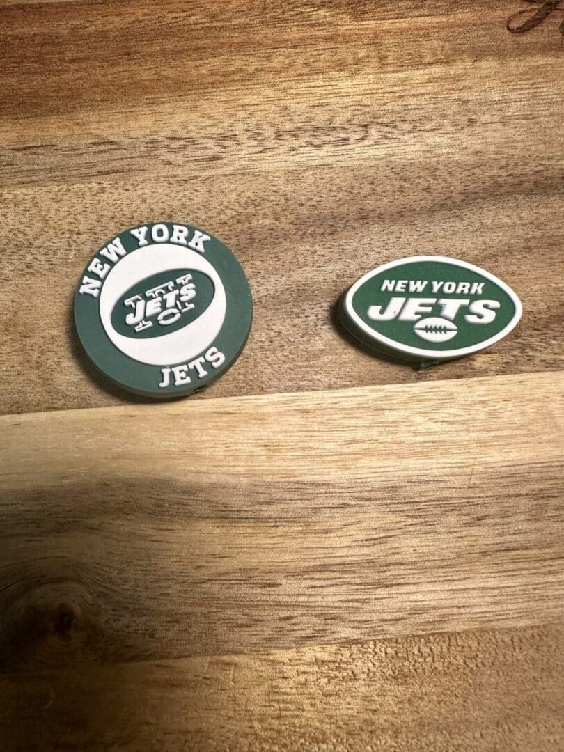 Two New York Jets accessories on a wooden surface. On the left is a circular NY football silicone focal bead with the team logo, and on the right is an oval-shaped NY football silicone focal bead with the same logo.