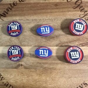 Six New York Giants silicone focal beads displayed on a wooden surface. The beads feature the team's logo and colors, with three circular designs and three football-shaped designs.