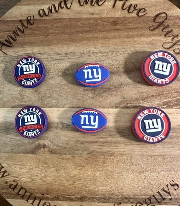 Six New York Giants silicone focal beads displayed on a wooden surface. The beads feature the team's logo and colors, with three circular designs and three football-shaped designs.