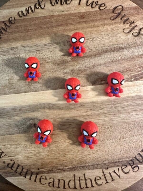 Six small 3D Spider-Man silicone focal beads are arranged on a wooden surface with engraved text around the edge.