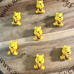 A wooden surface featuring seven small figurines of the "Winnie 3D Silicone Focal Bead" - a yellow bear holding a jar, arranged in a circular pattern. Text around the edge reads "Annie and the Five Guys" and includes a website URL.