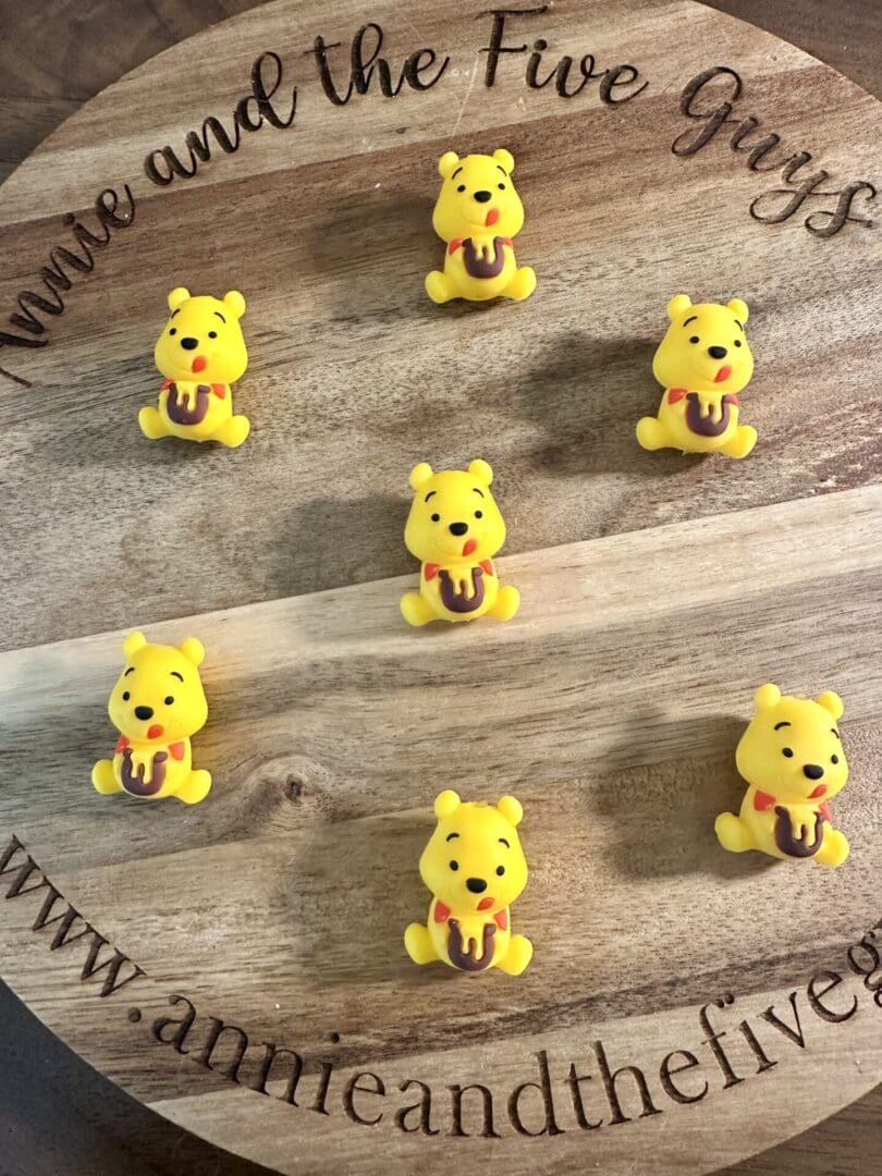 A wooden surface featuring seven small figurines of the "Winnie 3D Silicone Focal Bead" - a yellow bear holding a jar, arranged in a circular pattern. Text around the edge reads "Annie and the Five Guys" and includes a website URL.