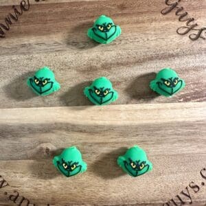 Six 3D Grinch silicone focal beads with expressive faces are arranged in two rows of three on a round wooden board engraved with text.