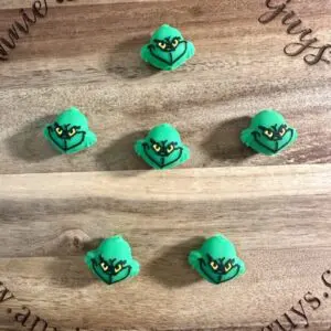 Six 3D Grinch silicone focal beads with expressive faces are arranged in two rows of three on a round wooden board engraved with text.