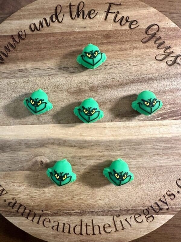 Six 3D Grinch silicone focal beads with expressive faces are arranged in two rows of three on a round wooden board engraved with text.