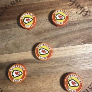 Five Kansas City Chiefs focal beads are arranged on a wooden surface with an engraved phrase partially visible.