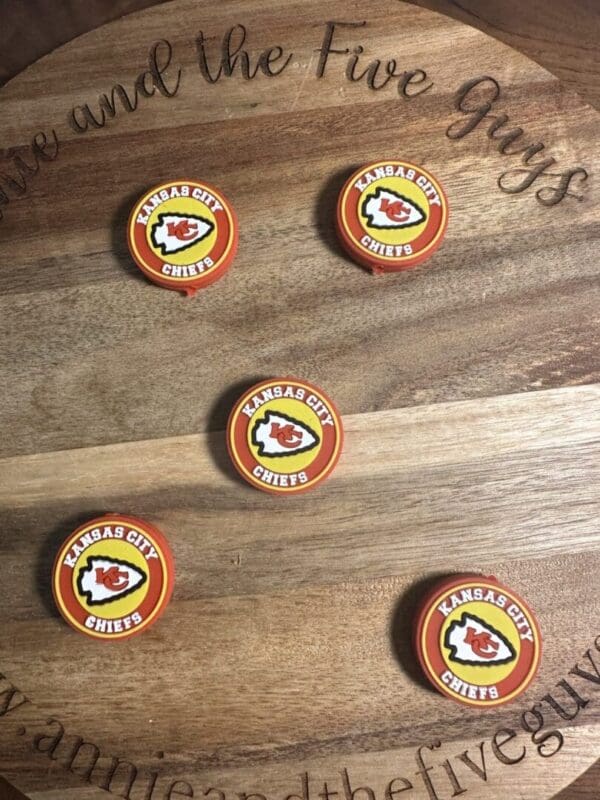 Five Kansas City Chiefs focal beads are arranged on a wooden surface with an engraved phrase partially visible.