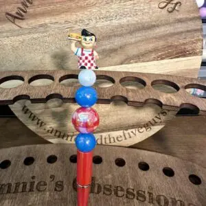 A small figurine of a man holding a sandwich tops a stack of beads on one of the 5 custom pens, standing on a wooden display that reads "annie and the five guys" and "annie's obsessions".