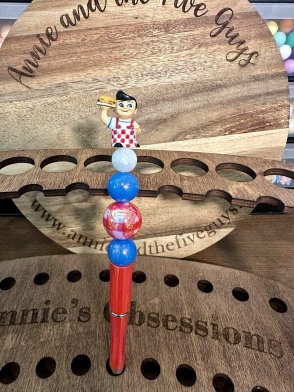 A small figurine of a man holding a sandwich tops a stack of beads on one of the 5 custom pens, standing on a wooden display that reads "annie and the five guys" and "annie's obsessions".