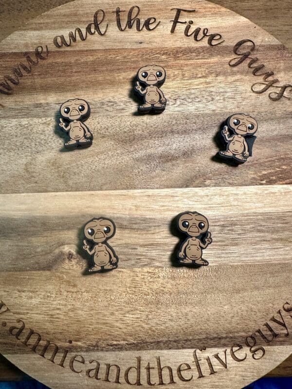 Five ET silicone focal beads featuring a character resembling an alien with a large head and big eyes are displayed on a wooden surface engraved with "annieandthefiveguys" and "Annie and the Five Guys.