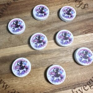 Round wooden board displaying eight circular silicone focal beads, each featuring a cartoon skeleton in a purple cloak holding pink flowers, named "Grim Reaper Riding a Unicorn.