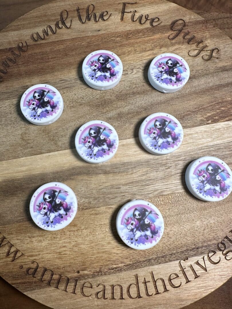 Round wooden board displaying eight circular silicone focal beads, each featuring a cartoon skeleton in a purple cloak holding pink flowers, named "Grim Reaper Riding a Unicorn.