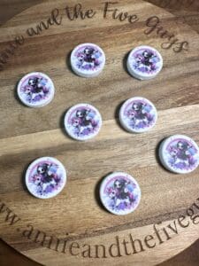 Wooden board displaying eight circular pins featuring the image of a grim reaper riding a unicorn, surrounded by the text "www.annieandthefiveguys.com".