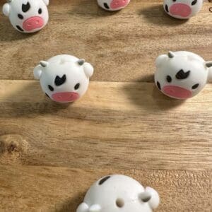 Six small, round Cow 3d silicone focal beads featuring pink snouts and black marks are arranged on a wooden surface.