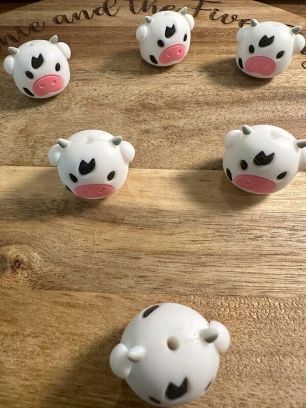 Six small, round Cow 3d silicone focal beads featuring pink snouts and black marks are arranged on a wooden surface.