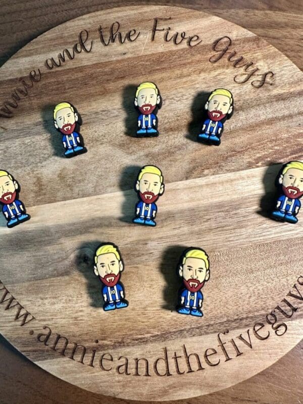 Round wooden board with the text "annie and the five guys" displaying nine small, illustrated Messi/soccer silicone focal beads depicting a bearded man with blond hair.