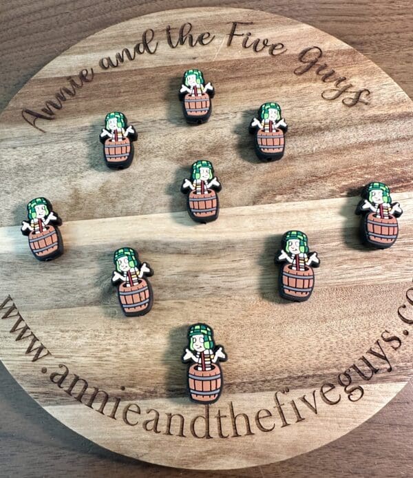 A circular wooden board with the text “Annie and the Five Guys” showcases nine El Chavo Silicone focal beads, depicting a character in a barrel with a green hat. The website www.annieandthefiveguys.com is also visible.