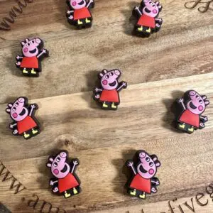 A wooden board showcases eight Peppa silicone focal beads, each designed as a small figurine of a cartoon pig dressed in a red dress, arranged in a circular pattern. The board is engraved with "annieandthefiveguys" and decorative text.