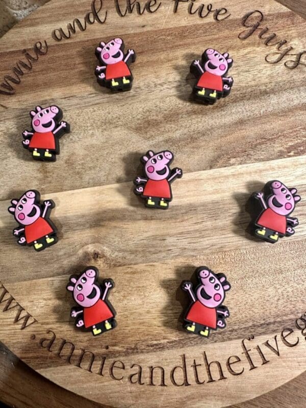 A wooden board showcases eight Peppa silicone focal beads, each designed as a small figurine of a cartoon pig dressed in a red dress, arranged in a circular pattern. The board is engraved with "annieandthefiveguys" and decorative text.