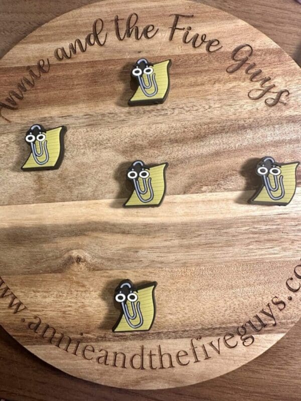 A round wooden board featuring five Paperclip and Paper silicone focal beads, each depicting a yellow cartoon character with glasses, and engraved with the text "annieandthefiveguys.com.