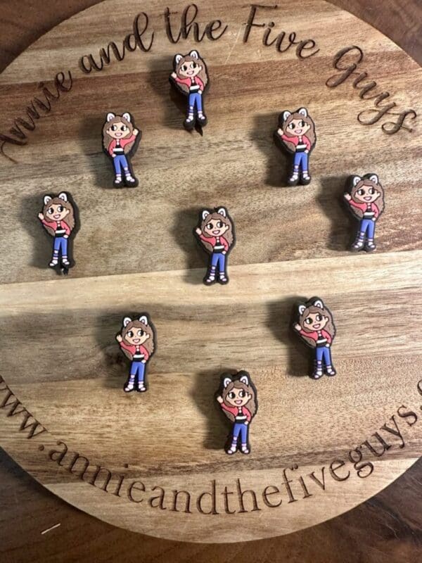 Nine small Gabby’s Dollhouse silicone focal beads, each featuring a character with cat ears, a blue shirt, and pink shoes, arranged on a round wooden surface.