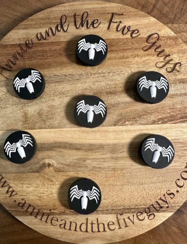 Round wooden board with the text "You're and the Five Guys" and a website at the bottom. Seven Venom silicone focal beads, featuring white spider logos, are arranged on the board.