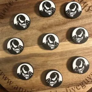 A wooden board displays nine black and white circular tokens featuring a stylized skull image, each labeled as a Venom face silicone focal bead. The board is inscribed with "Annie and the Five Guys" and a website link.