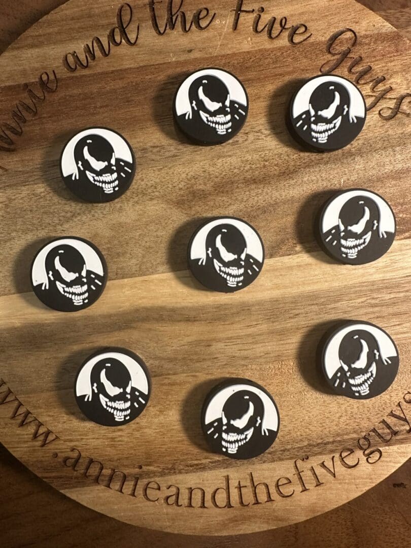 A wooden board displays nine black and white circular tokens featuring a stylized skull image, each labeled as a Venom face silicone focal bead. The board is inscribed with "Annie and the Five Guys" and a website link.