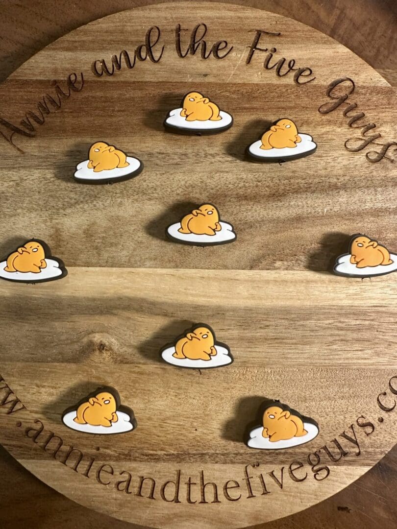 Wooden board displaying eight silicone focal beads depicting Gudetama, a resting yellow creature on a white cloud, arranged in a grid pattern. Text around the edges reads "Annie and the Five Guys" and a website address.