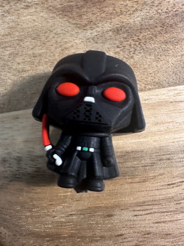 A 3D Darth Vader silicone focal bead, depicting a character in black armor and helmet with red eyes and wielding a red lightsaber, standing on a wooden surface.