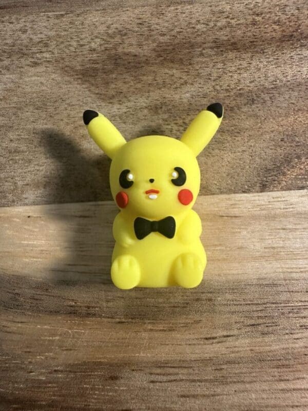 A small Pocket Monster Silicone Focal Bead 3D figurine of Pikachu with a black bowtie, sitting on a wooden surface.