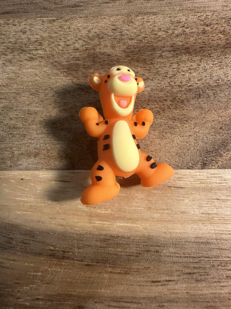 A 3D Tigger silicone focal bead, shaped like an orange tiger with black stripes and a cheerful expression, is standing on a wooden surface.