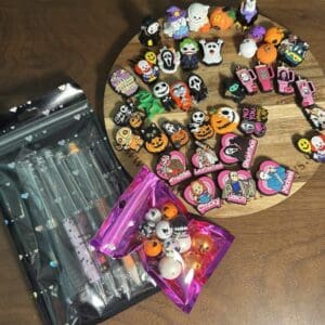 A wooden table showcases the Halloween Bundle, which includes an assortment of rubber charms and pins inspired by Halloween. To the left, two packages feature clear plastic pens and small balls, all adorned with Halloween themes.