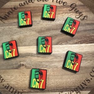 A circular wooden surface with six square Soccer silicone focal beads featuring colorful graphics of a green and red figure with "CHZ" text. The beads are arranged randomly. The surface reads "Annie and the Five Guys" and a web address.