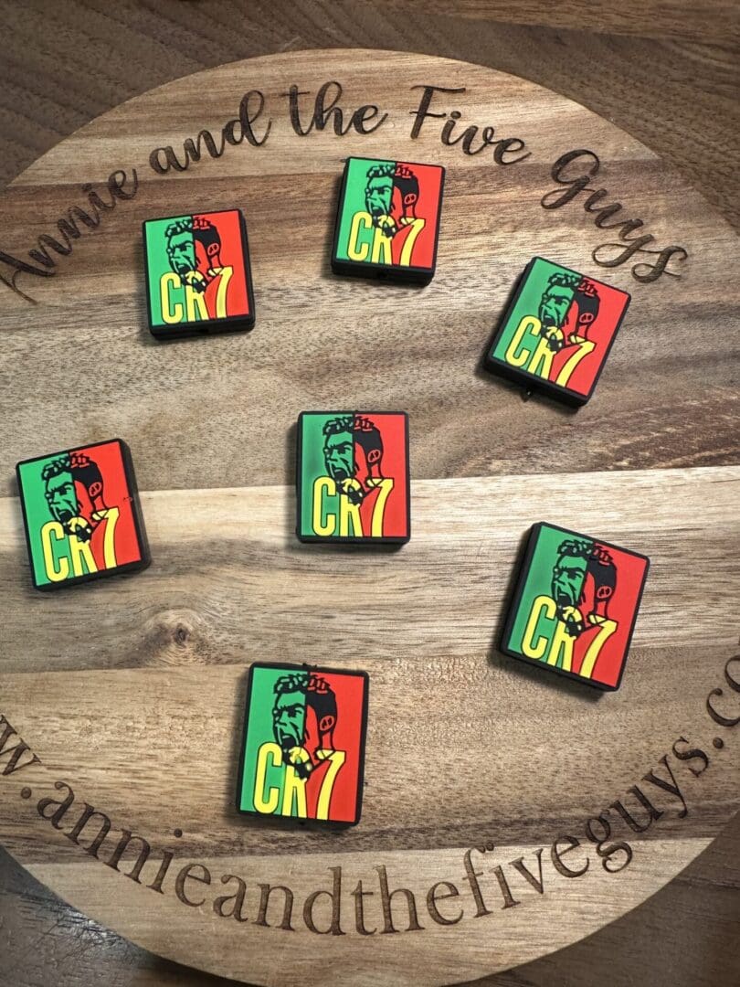 A circular wooden surface with six square Soccer silicone focal beads featuring colorful graphics of a green and red figure with "CHZ" text. The beads are arranged randomly. The surface reads "Annie and the Five Guys" and a web address.