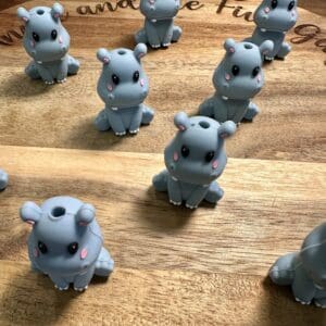 Several Hippopotamus silicone focal beads, featuring a small gray design with pink cheeks, are arranged on a wooden surface.