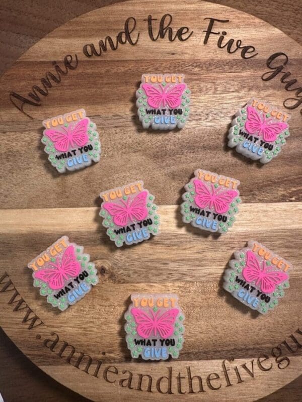 Decorated cookies with pink butterfly designs, featuring the phrase "You get what you give" on a wooden tray, accompanied by a "You get what you give" silicone focal bead.