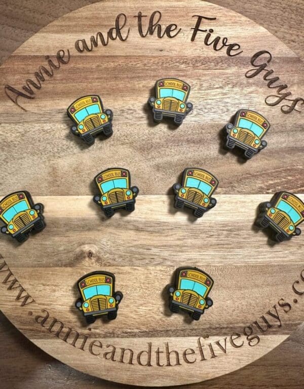 A wooden surface with a circular "Annie and the Five Guys" logo. Nine Magic School Bus silicone focal beads are arranged in a grid.