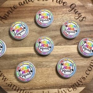 Round wooden board displaying several "Lunch Hero Squad" silicone focal beads with colorful designs. The board is engraved with the text "Annie and the Five Guys www.annieandthefiveguys.com.