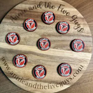 A wooden board displaying nine round coasters, each featuring an Atlanta football silicone focal bead with the Atlanta Falcons logo. The board is inscribed with "Annie and the Five Guys" and a website URL.