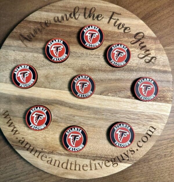 A wooden board displaying nine round coasters, each featuring an Atlanta football silicone focal bead with the Atlanta Falcons logo. The board is inscribed with "Annie and the Five Guys" and a website URL.