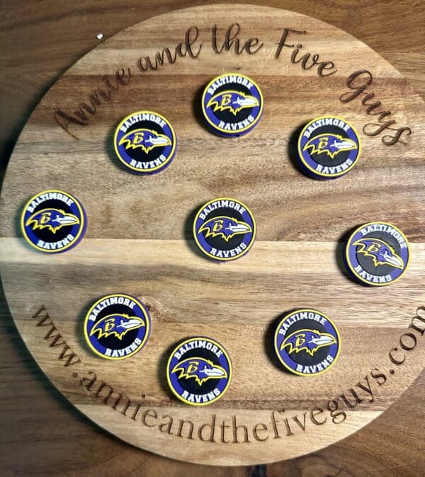 A wooden board with the words "Annie and the Five Guys" displaying eight Baltimore Football Silicone Focal Beads, each featuring the Ravens logo.