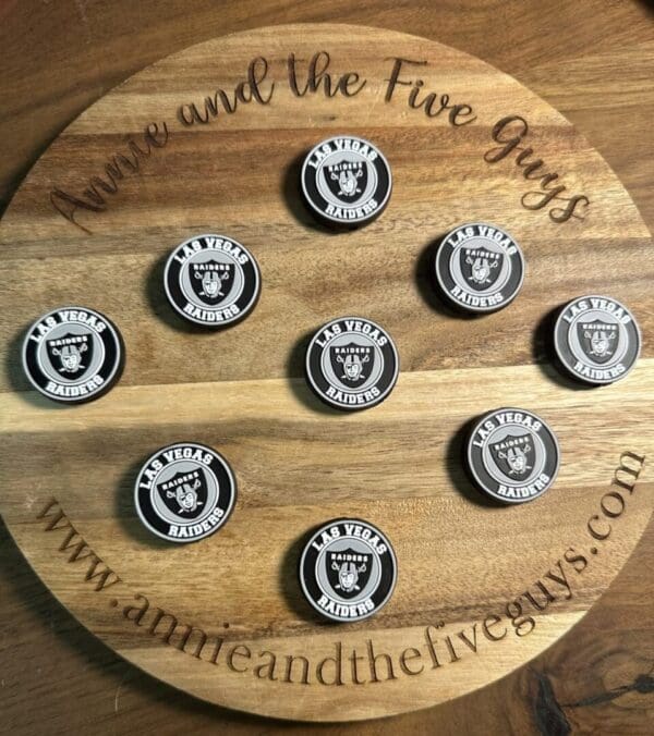 A round wooden board with "Annie and the Five Guys" text displays nine Las Vegas football silicone focal beads, arranged in a grid. The website "www.annieandthefiveguys.com" is visible on the board.