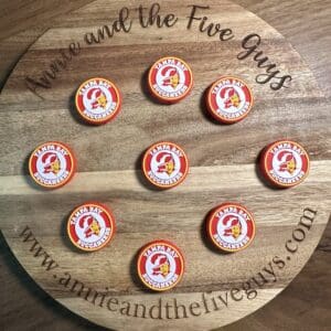 A wooden board displays nine silicone focal beads featuring the Tampa Bay Buccaneers logo, with the text "Annie and the Five Guys" and a website URL circling the perimeter.