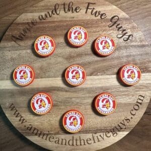 A wooden board displays nine silicone focal beads featuring the Tampa Bay Buccaneers logo, with the text "Annie and the Five Guys" and a website URL circling the perimeter.