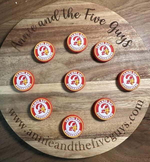 A wooden board displays nine silicone focal beads featuring the Tampa Bay Buccaneers logo, with the text "Annie and the Five Guys" and a website URL circling the perimeter.